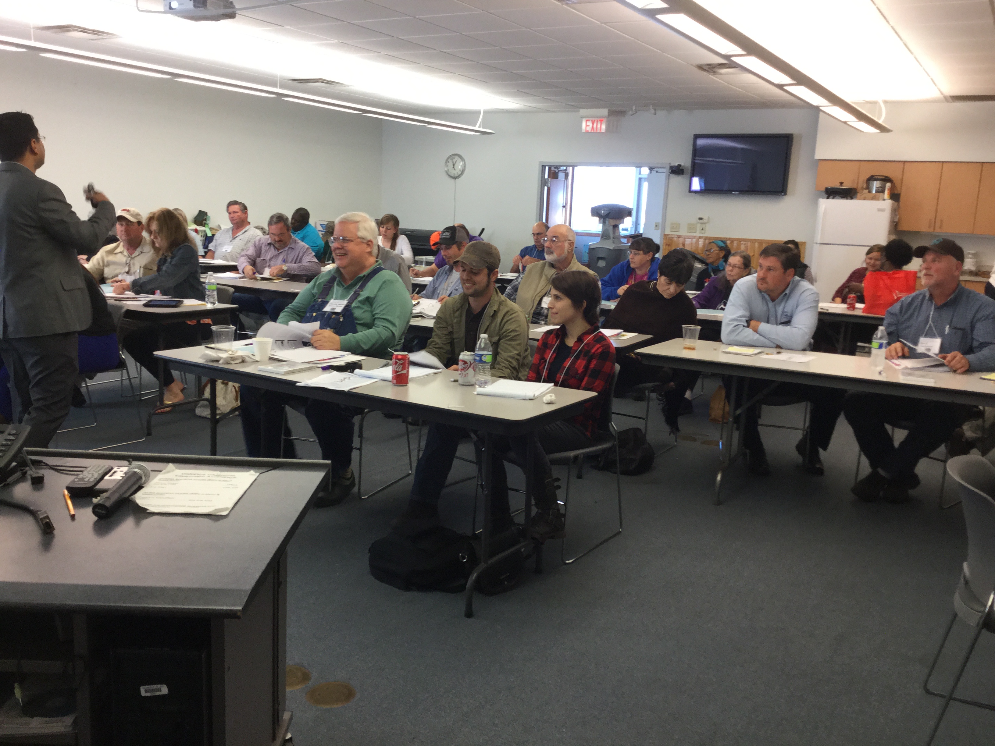 In a classroom or seminar setting, a speaker stands at the front of the room addressing a group of attendees seated at tables. The participants appear engaged, with some taking notes and others listening attentively. The room is well-lit with fluorescent lights and features a wall-mounted television, a clock, and a whiteboard. Tables are equipped with papers, notebooks, and drinks, indicating a professional or educational environment such as a workshop or training session.